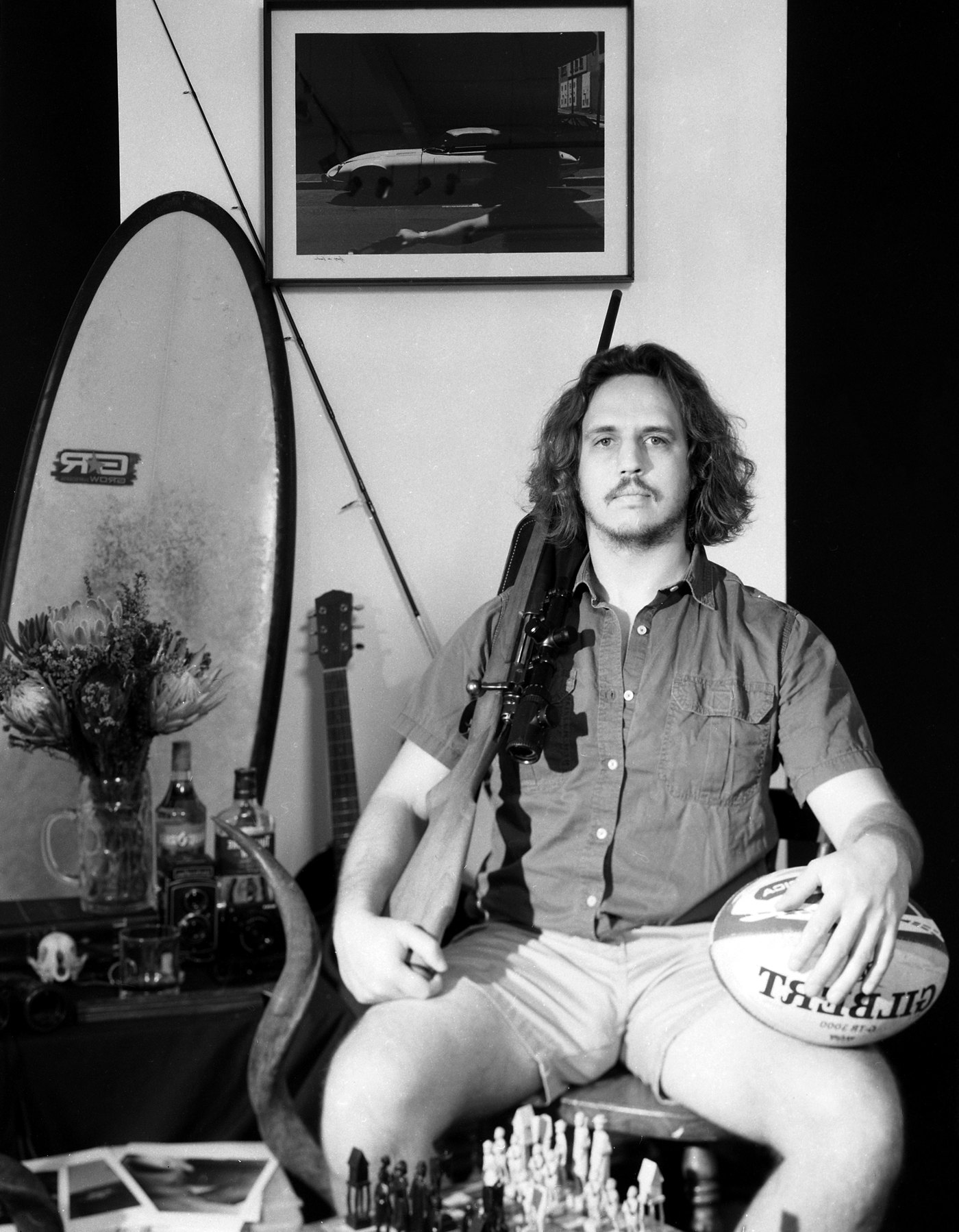A finished 4x5 black and white portrait of a seated man with a surfboard, guitar and chessboard behind him