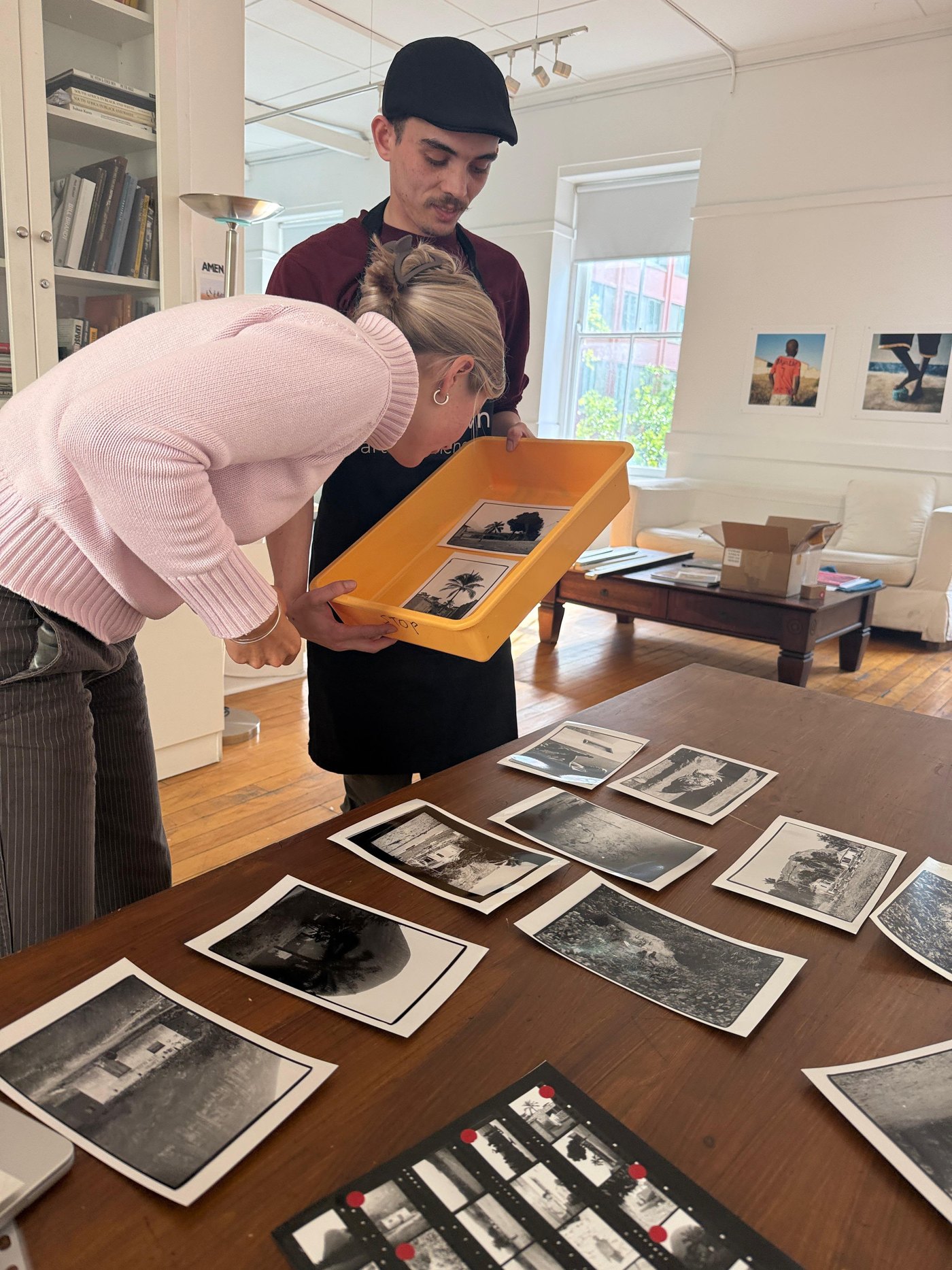 A woman and a man in an apron looking over finished prints in a yellow tray, with more prints and a marked contact sheet laid out on the table