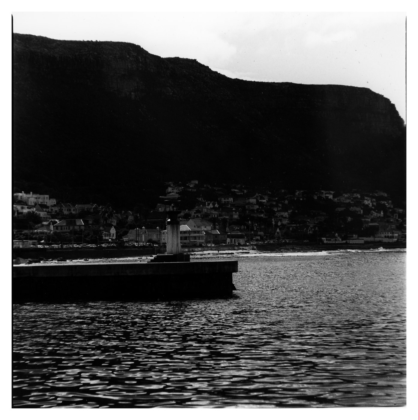 A black and white print of a harbour wall and lighthouse below a dark cliff