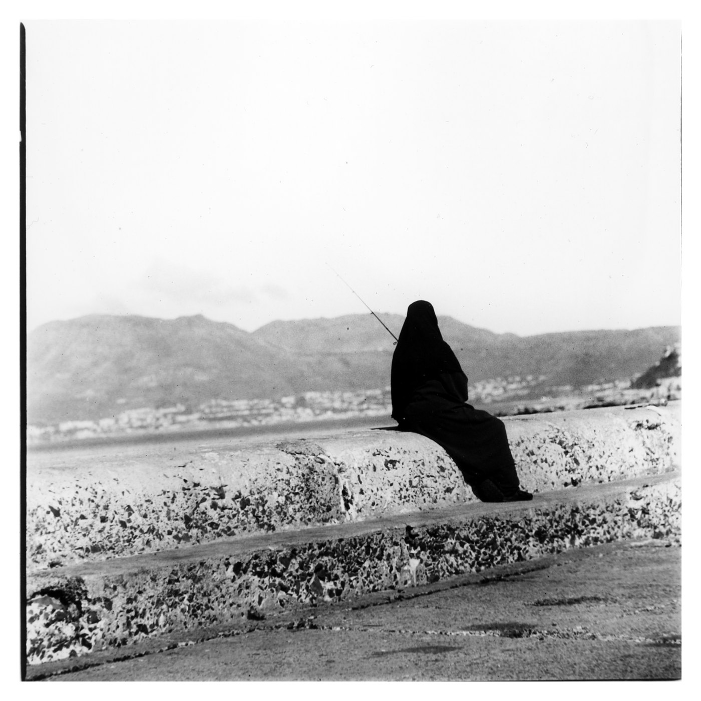 A black and white print of a figure in dark robes sitting on a sea wall above a bay