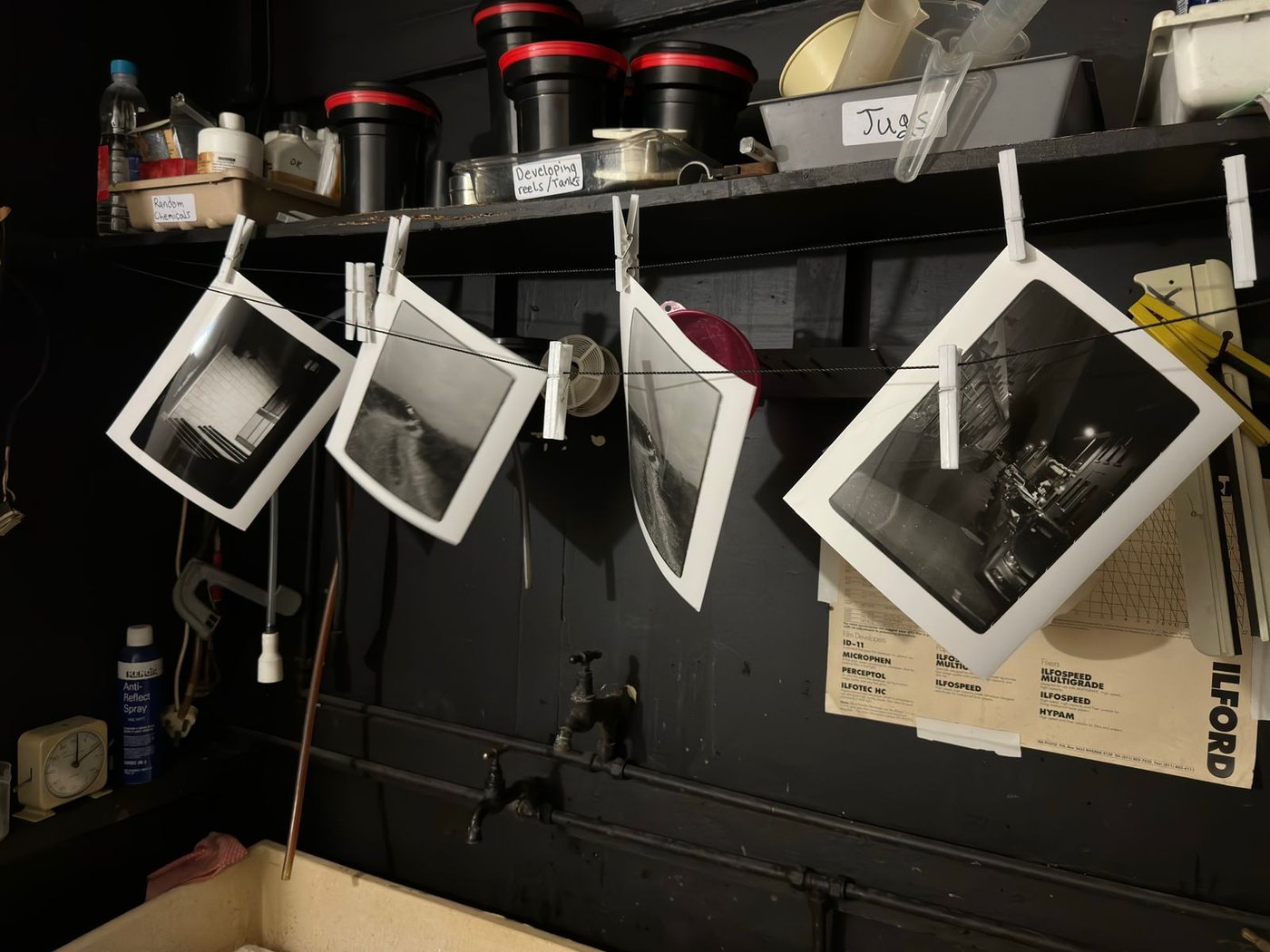 Four freshly made black and white prints hanging from pegs on a line in the darkroom, with shelves of developing tanks above